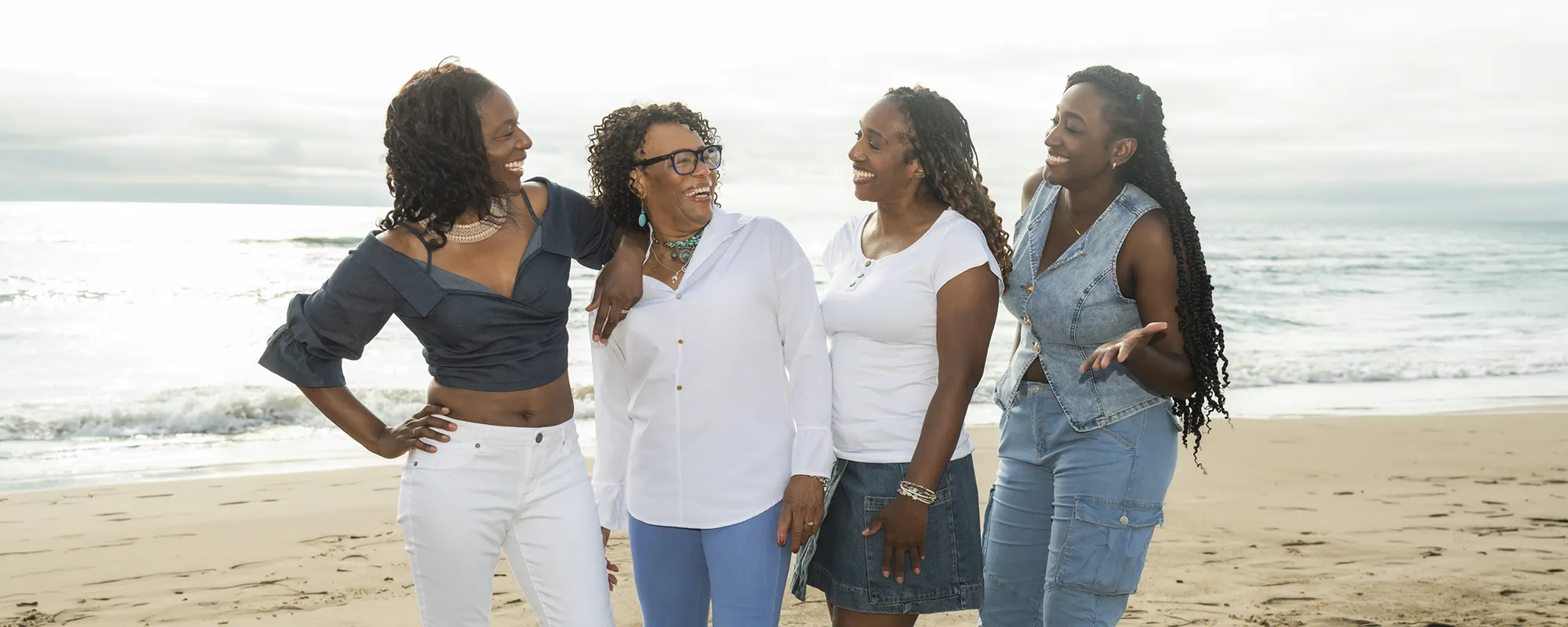Four women laughing together on a sandy beach beside the water