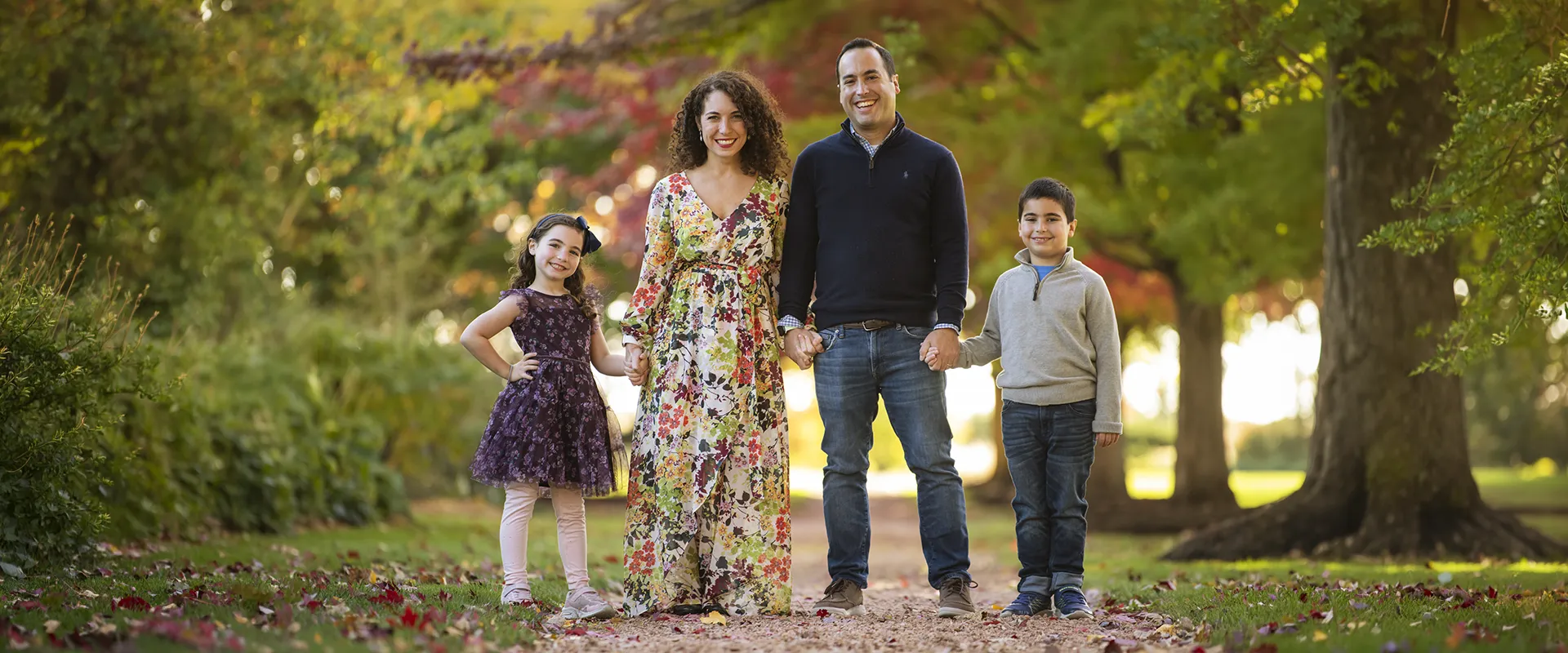 Family of four holding hands on a park path surrounded by autumn foliage