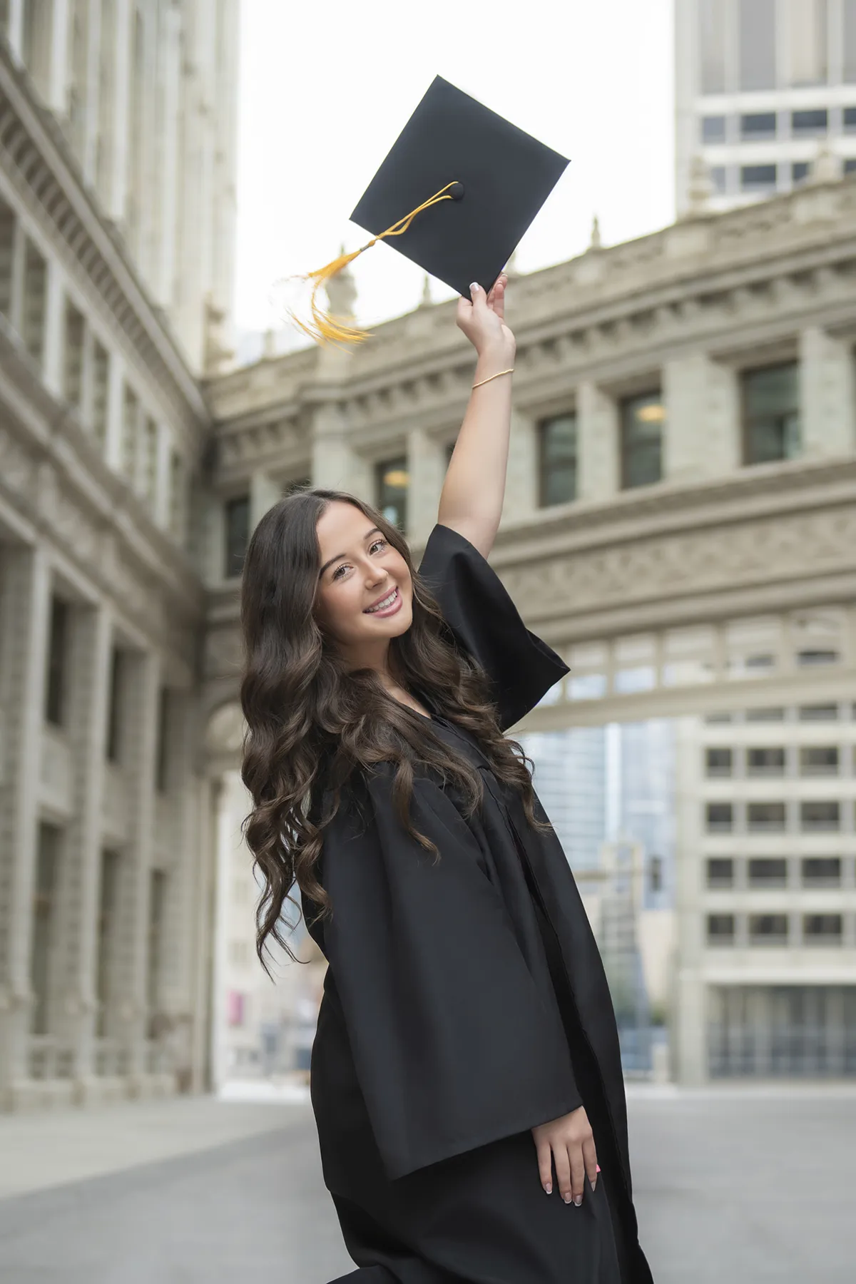 Graduate smiling and holding her graduation cap overhead among city buildings