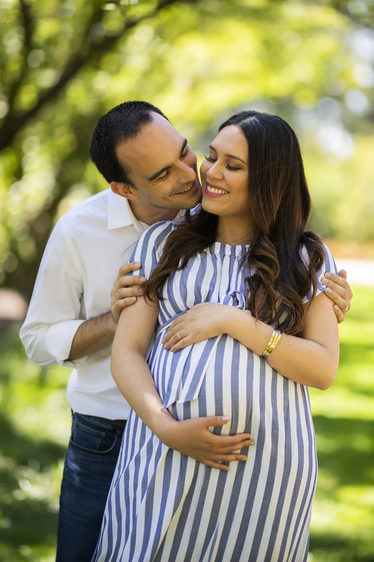 Expectant mother and her partner embracing outdoors beneath leafy trees